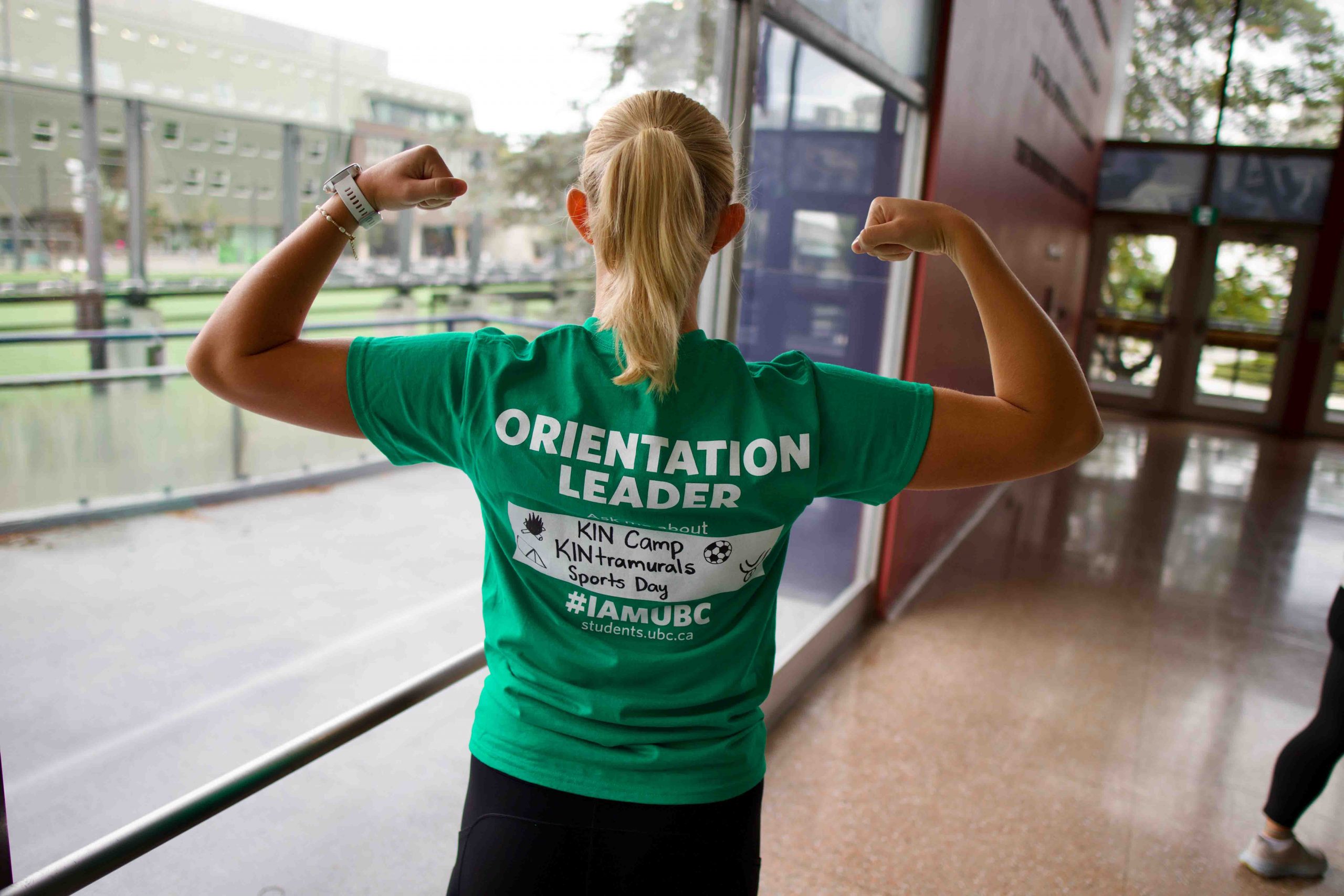 Kinesiology student facing away from the camera to show the back of her t-shirt that says "Orientation Leader". She is posing with her arms flexed in fists by her ears.