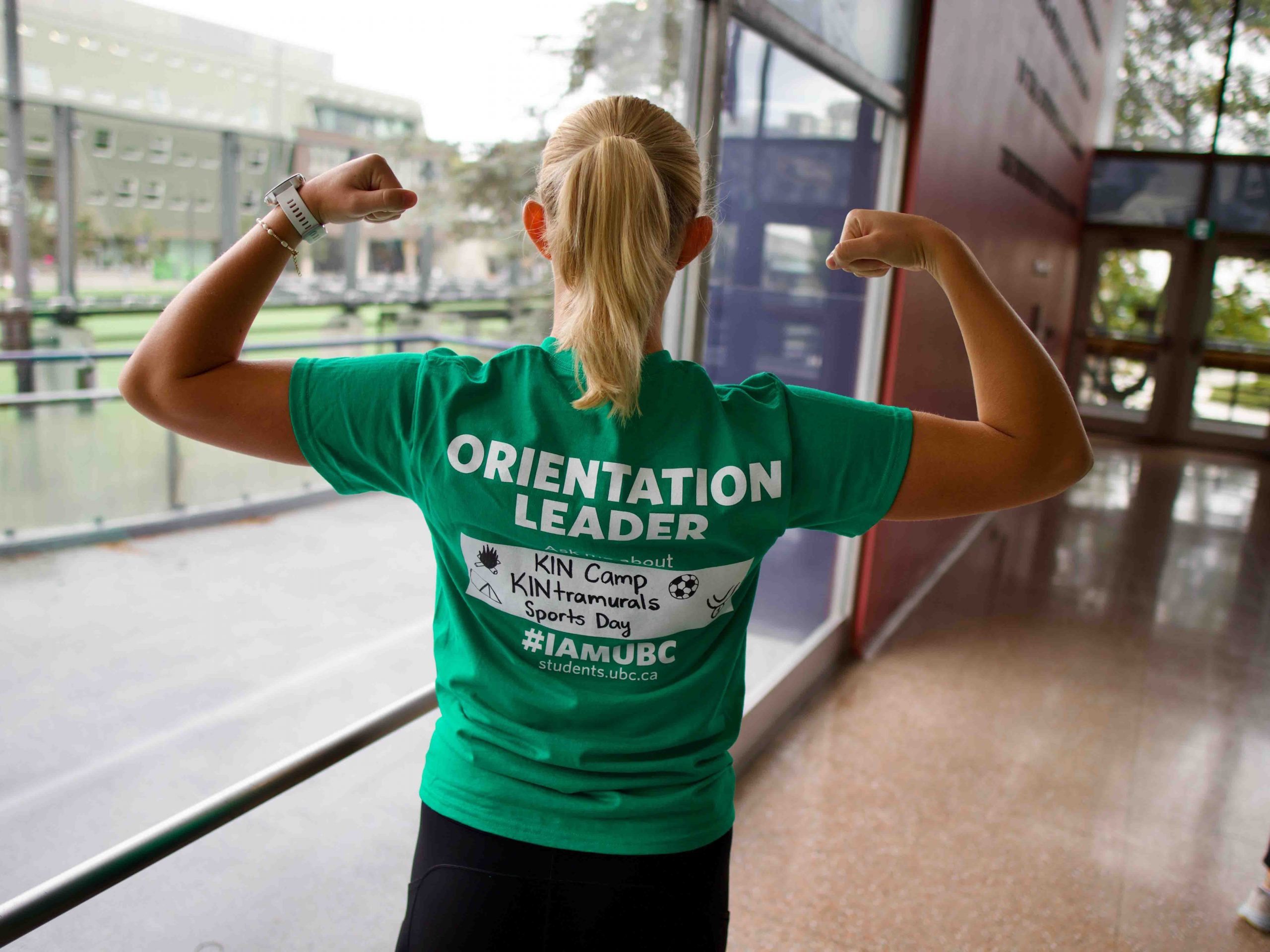 Kinesiology student facing away from the camera to show the back of her t-shirt that says "Orientation Leader". She is posing with her arms flexed in fists by her ears.
